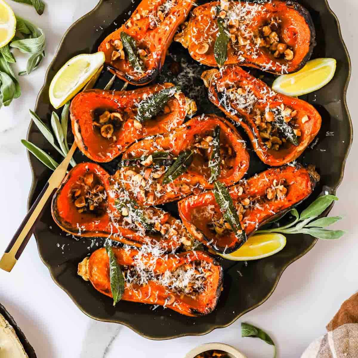 Roasted butternut squash halves topped with nuts, herbs, and grated cheese on a platter with lemon wedges.